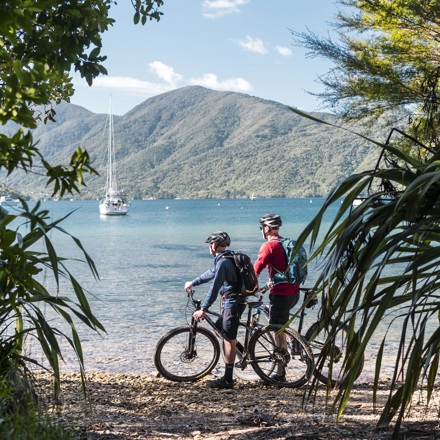 Two male bikes parked on the beach just off the Queen Charlotte Track with a yacht and the Marlborough Sounds behind.