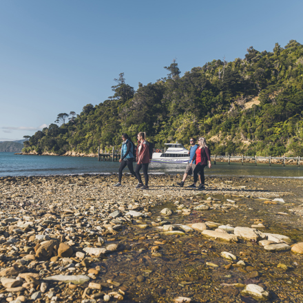 Four people walk along the beach at Ship Cove/Meretoto with a Cougar Line boat in the background.