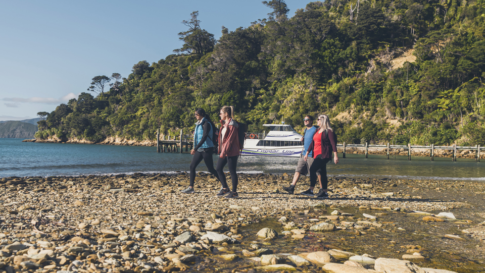 Four people walk along the beach at Ship Cove/Meretoto with a Cougar Line boat in the background.