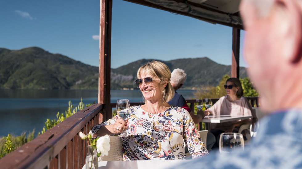 A woman with glass of wine sits on balcony with partner and couple behind, and view of Marlborough Sounds at Punga Cove