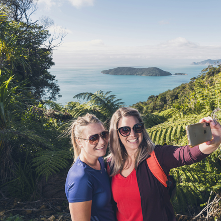 Two women take selfies at the lookout with Motuara Island in the background on the Queen Charlotte Track.