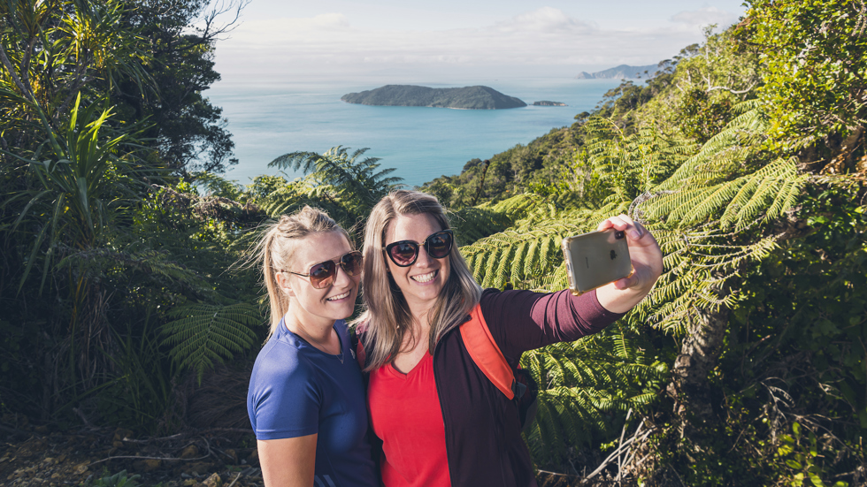 Two women take selfies at the lookout with Motuara Island in the background on the Queen Charlotte Track.